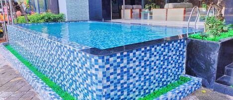 A swimming pool with blue and white mosaic tiles, surrounded by a green border and a tiled walkway.