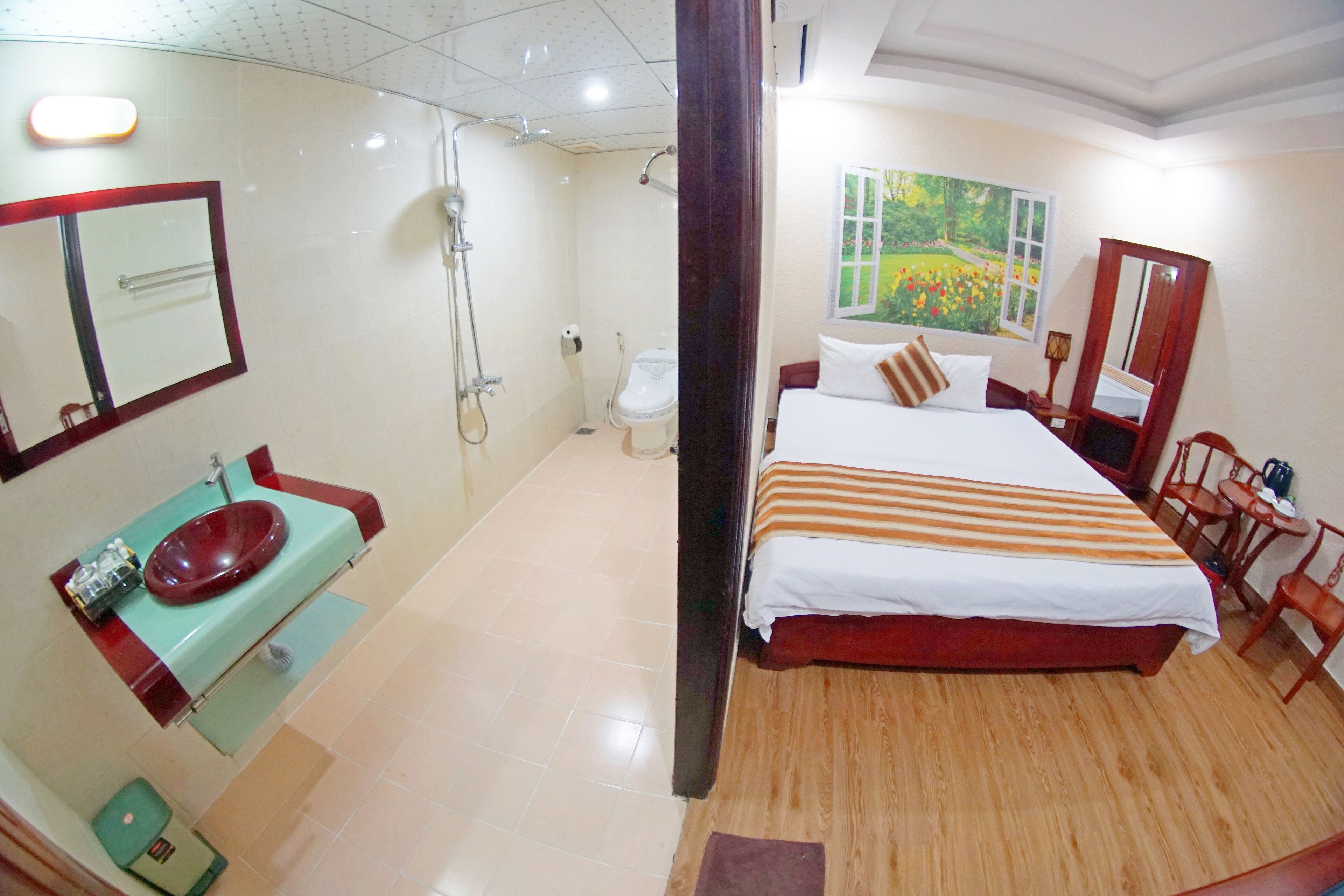 A hotel room with a large bed, a wooden floor, a bathroom with a sink and toilet, and a painting of a garden on the wall.
