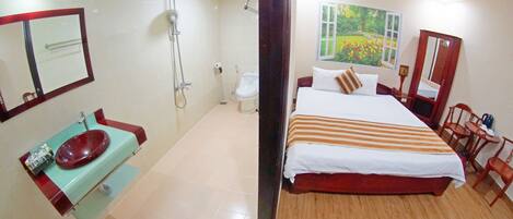 A hotel room with a large bed, a wooden floor, a bathroom with a sink and toilet, and a painting of a garden on the wall.