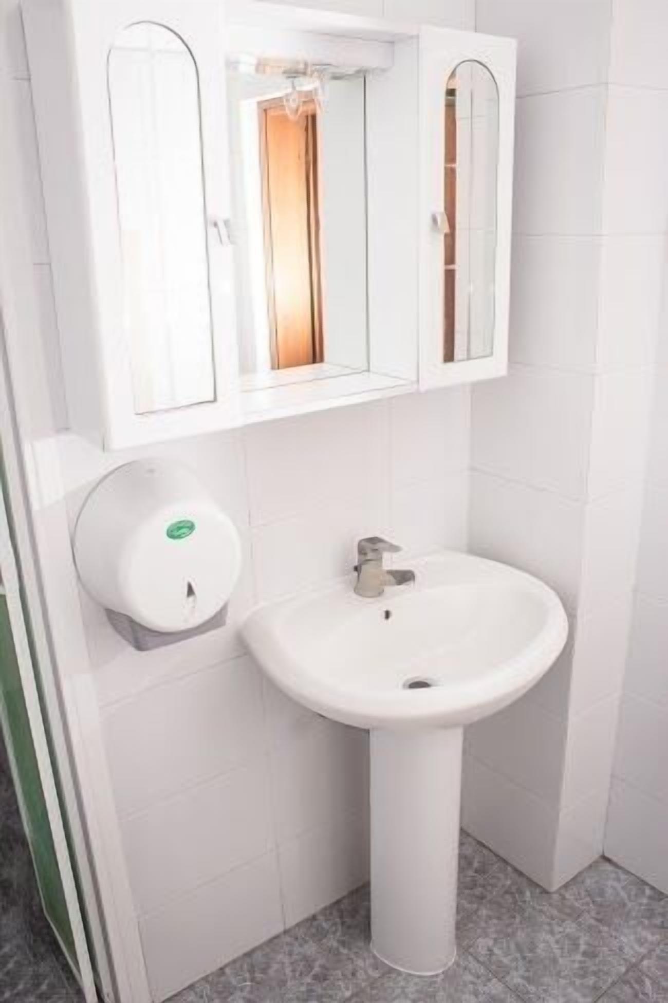 A white bathroom with a pedestal sink, a mirror above the sink, and a soap dispenser on the wall.