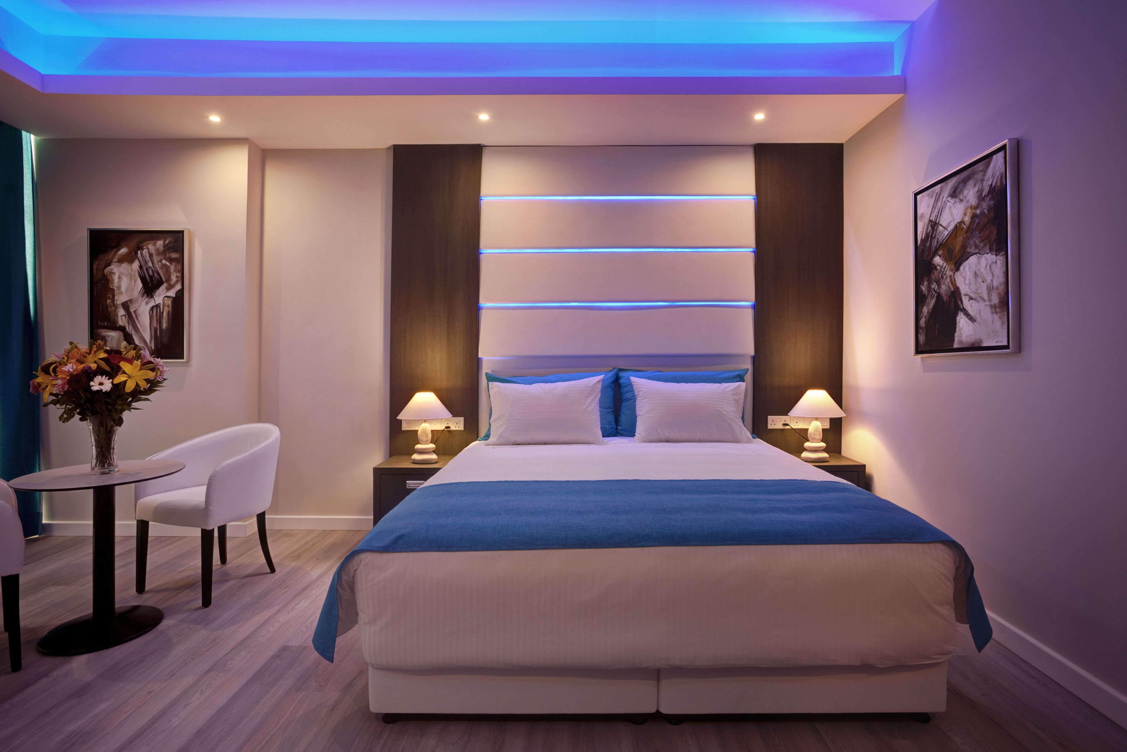 A modern hotel bedroom with a large bed, bedside tables with lamps, a small round table with chairs, and a vase of flowers.