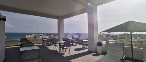 A rooftop terrace with tables and chairs, a sun umbrella, and a view of the sea.