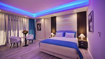 A modern hotel room with a large bed, two bedside tables with lamps, a small table with flowers, and a seating area with two chairs.