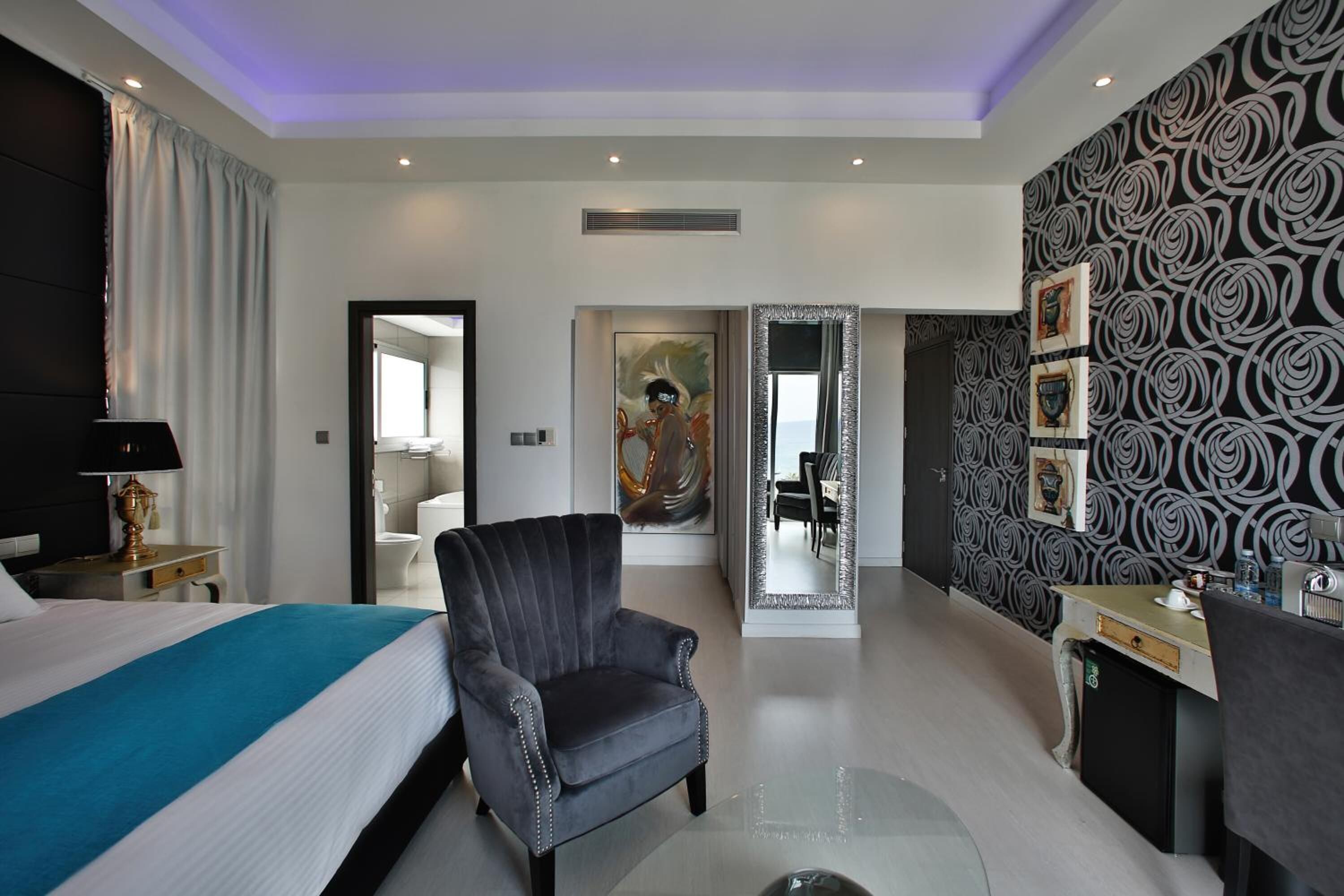 A modern hotel room with a large bed, a sitting area with a chair, a desk, and a bathroom visible through an open door.