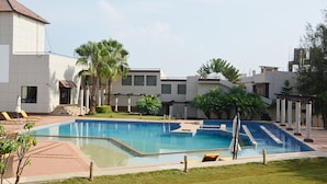 A swimming pool with a sunbathing area, palm trees, and a modern building with a tiled facade.