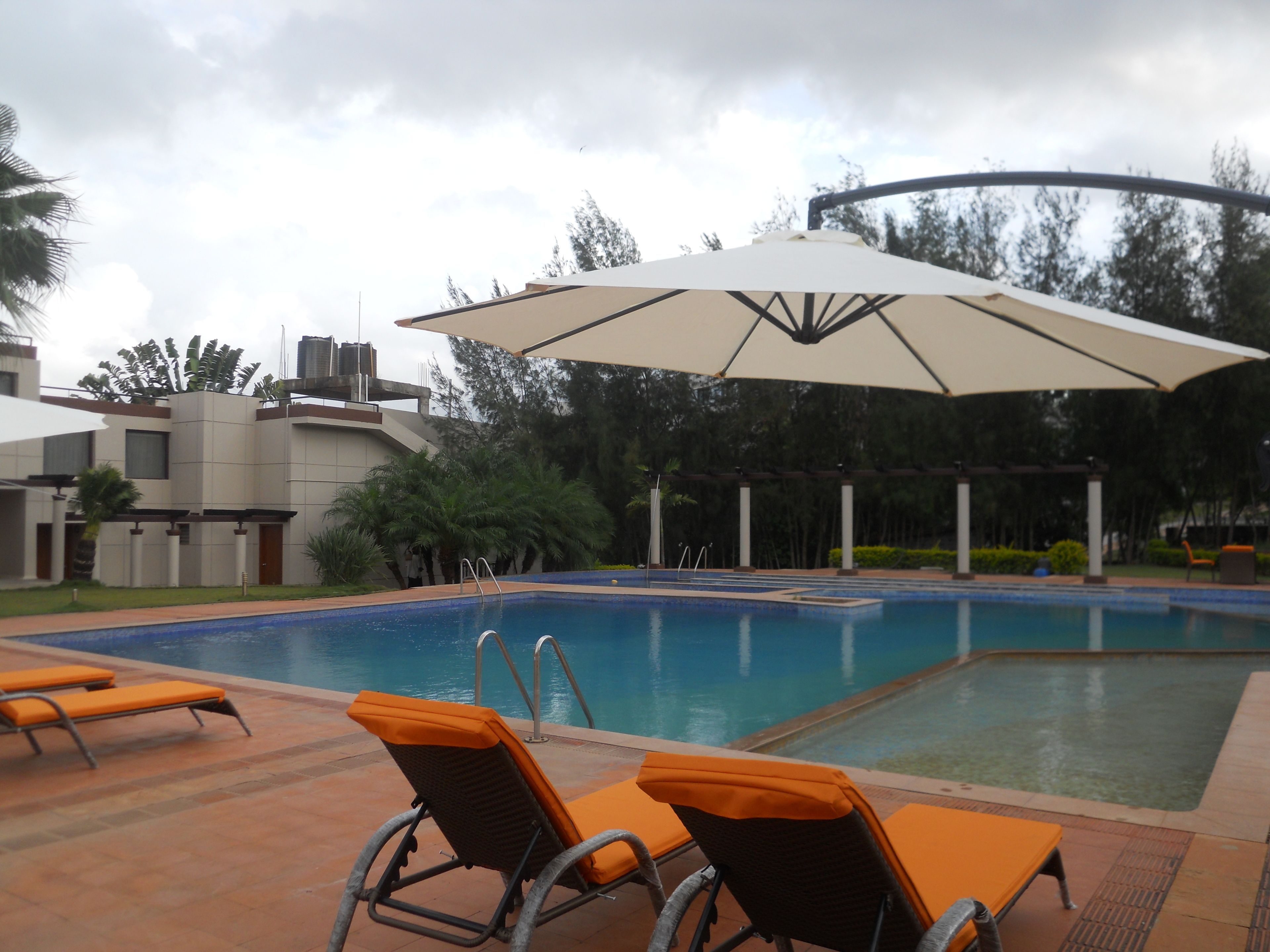 Piscina con sillones de descanso y sombrillas, rodeada de vegetación y edificios modernos.