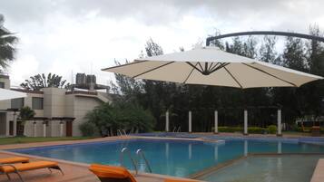 Piscina con sillones de descanso y sombrillas, rodeada de vegetación y edificios modernos.