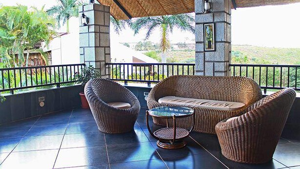 A covered outdoor seating area with wicker furniture and a small table, overlooking a lush landscape with palm trees.