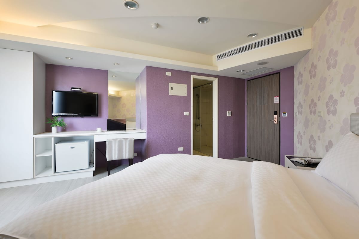 A modern hotel room with a bed, a desk, a television, and a shower area.