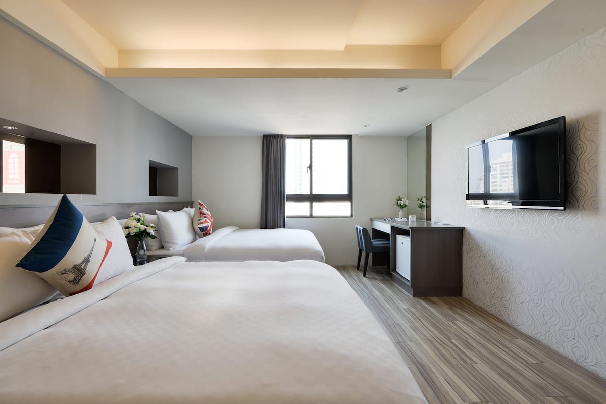 A hotel room with two beds, a desk, a TV, and a window.