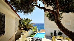 A pool area with lounge chairs, a view of the sea, and a building with a tiled roof.