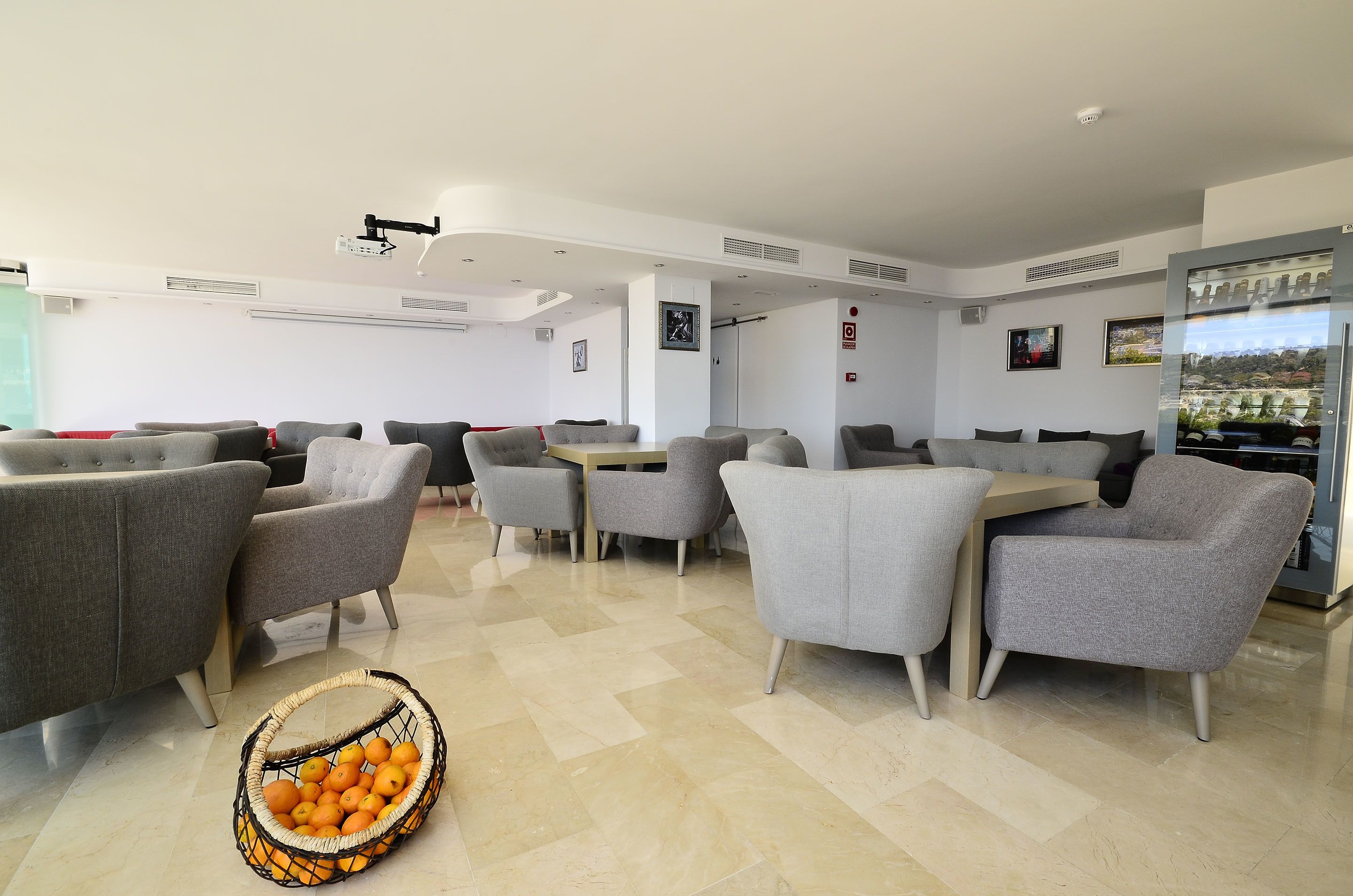 A modern hotel lobby with grey upholstered seating, a central table, and a basket of oranges on the floor.