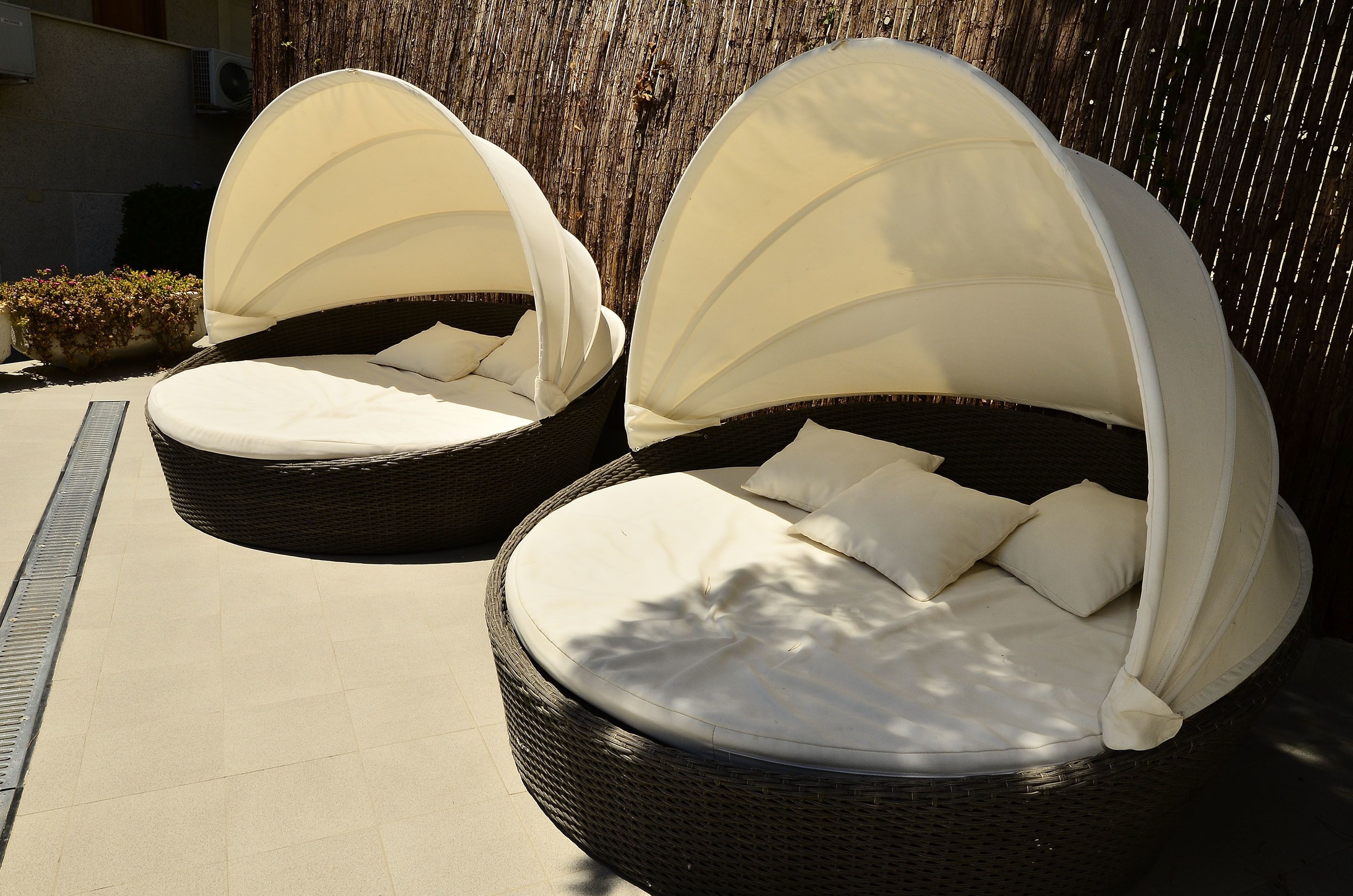 Deux fauteuils de jardin en osier, dotés de coussins blancs et de toiles de protection, sont placés sur une terrasse carrelée.