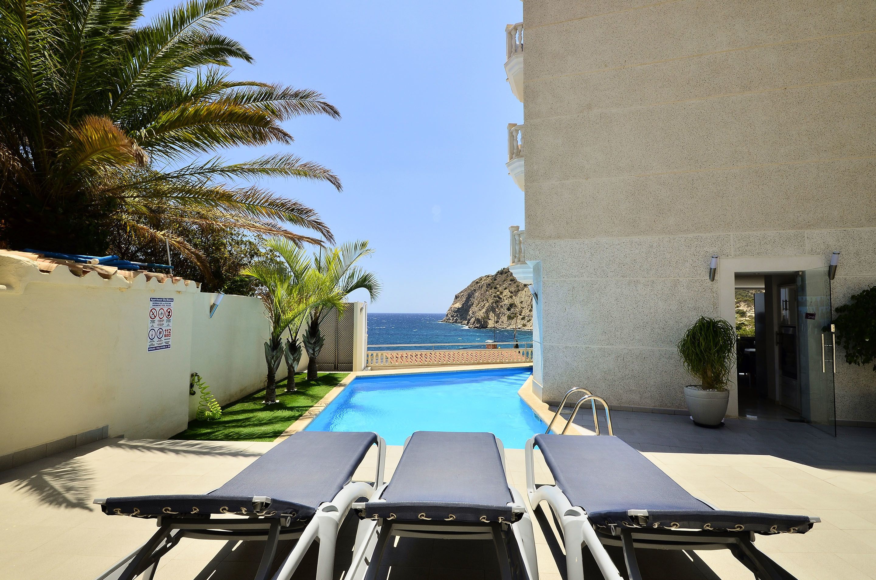 Un área de piscina con sillones, vista al mar y un edificio de fachada de piedra.