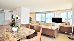 A modern living room with a large window, a flat-screen TV, and a dining area with a marble table.