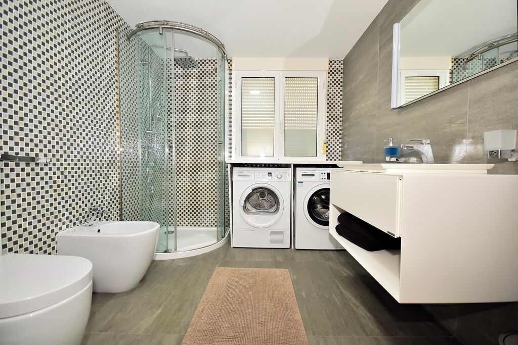 Un baño moderno con mampara de vidrio, lavadora, inodoro y lavamanos.