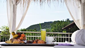 A breakfast setup with fruits, pastries, and a glass of juice on a tray, under a canopy with a view of a green hill. - Studios & Apartments Efrosini (Dio-Olympos)