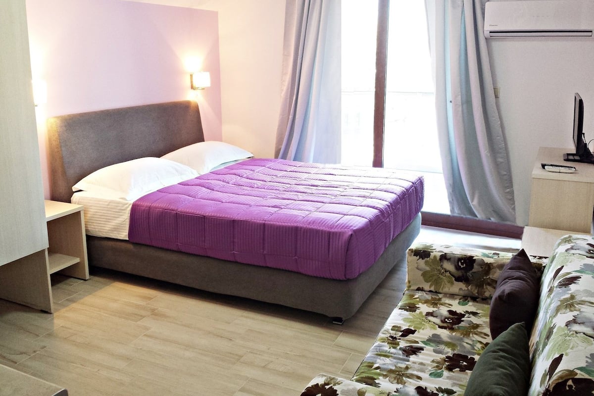 A bedroom with a bed, a nightstand, a desk, a chair, and a window with curtains.
