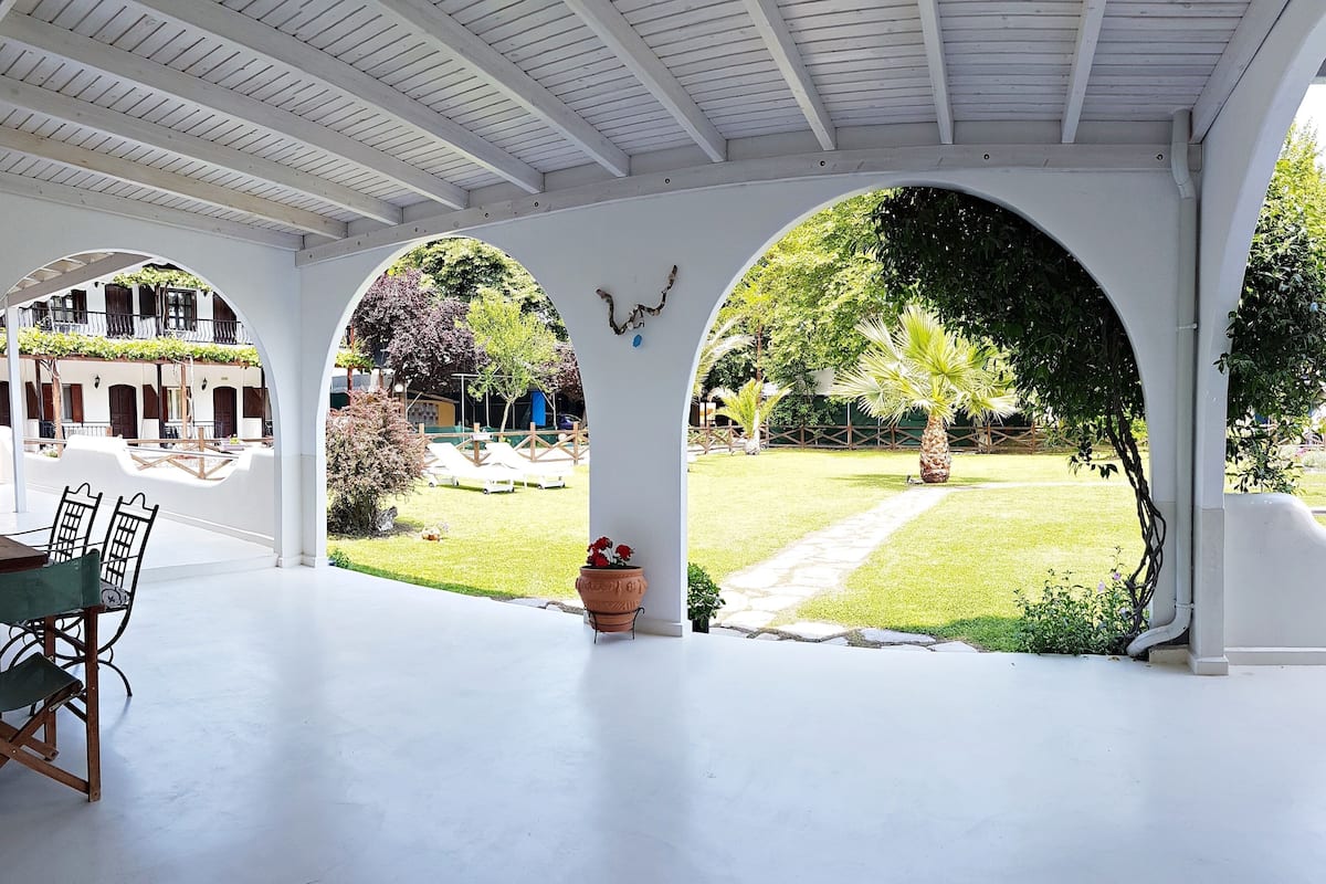 A covered patio area with white columns and arches, a seating area with chairs, and a view of a well-maintained garden with a lawn and trees.