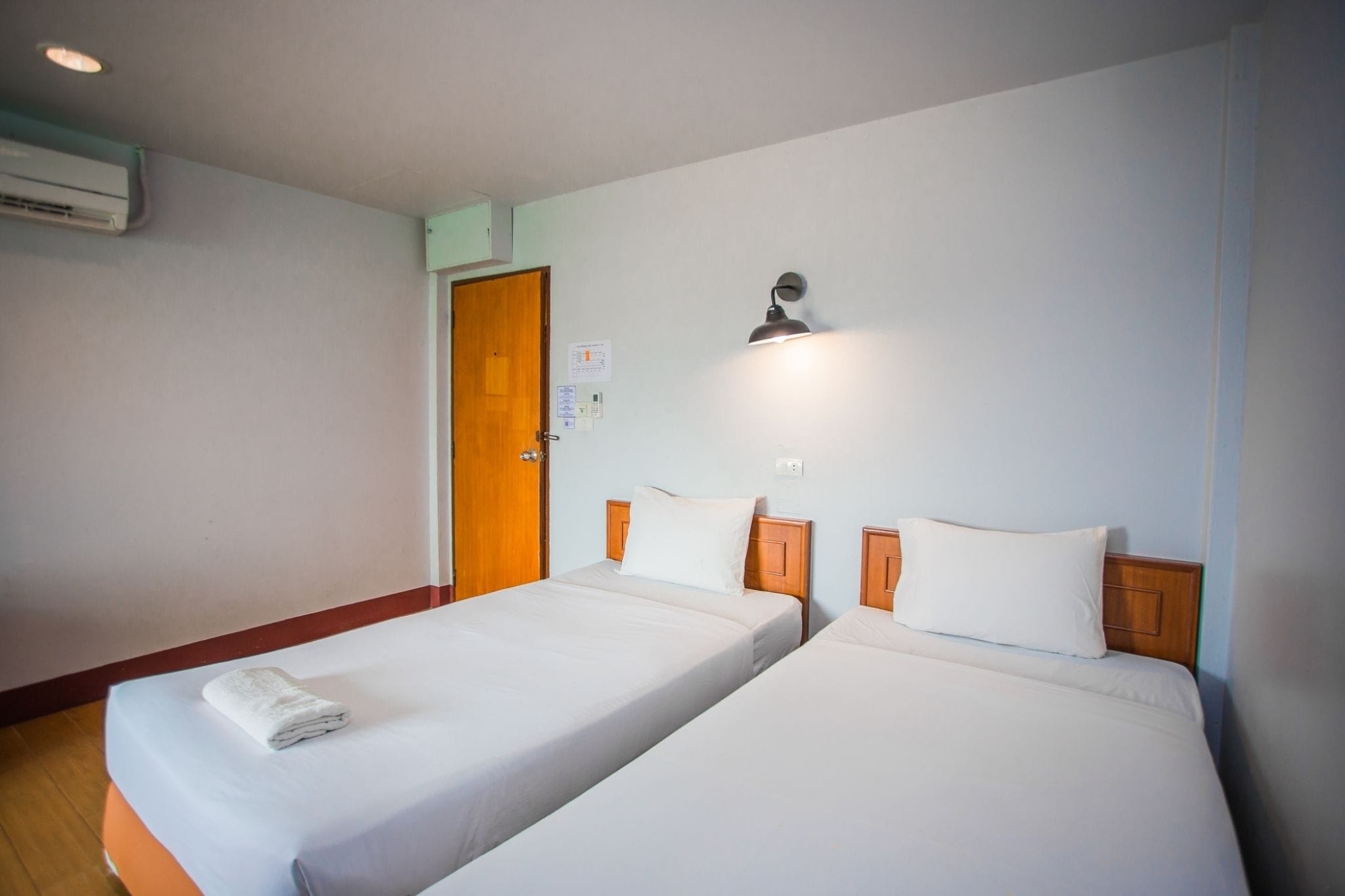 A hotel room with two single beds, a wooden door, an air conditioning unit, and a wall-mounted light fixture.