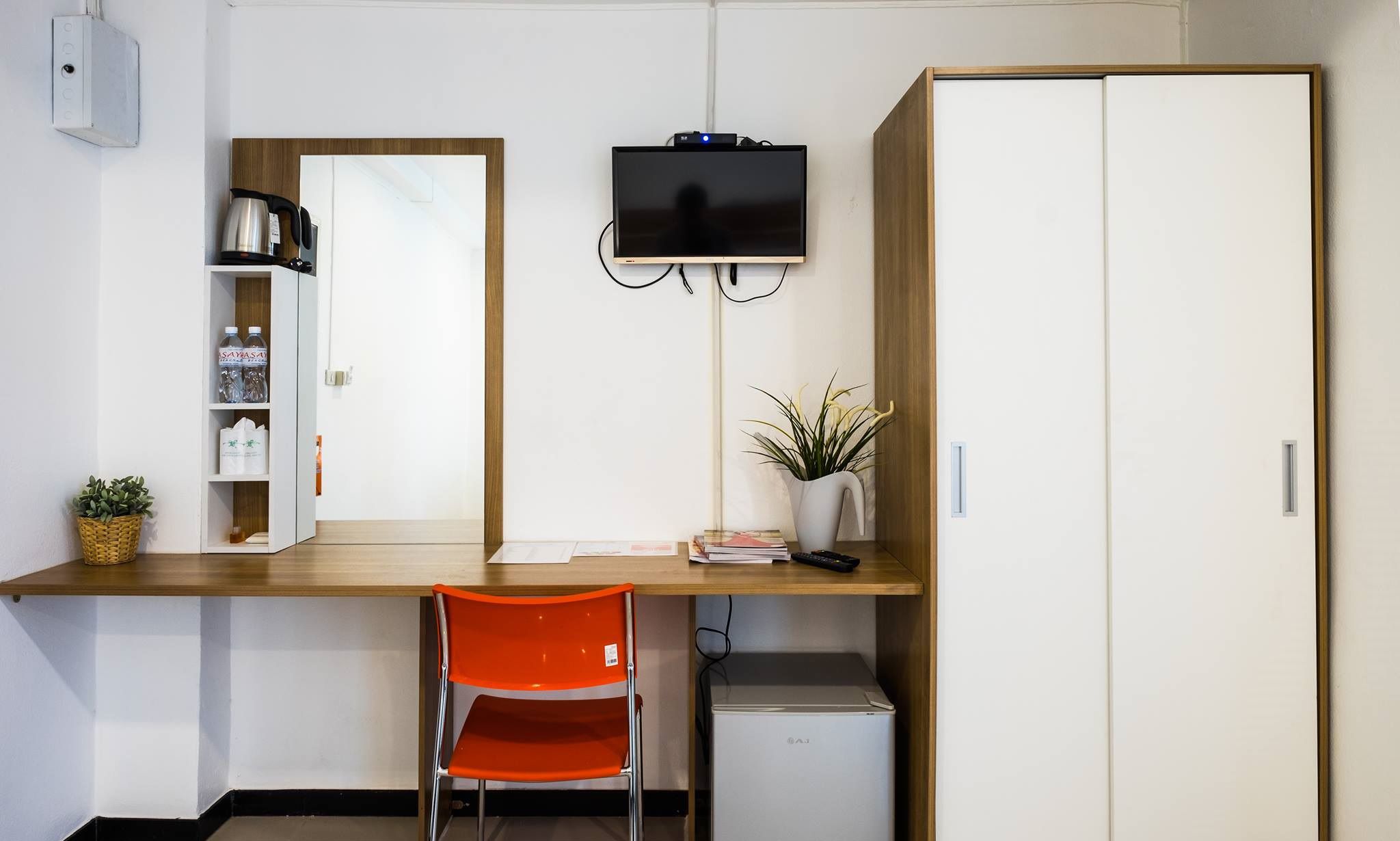 A compact room with a wooden desk, a television mounted on the wall, a small refrigerator, and a chair.
