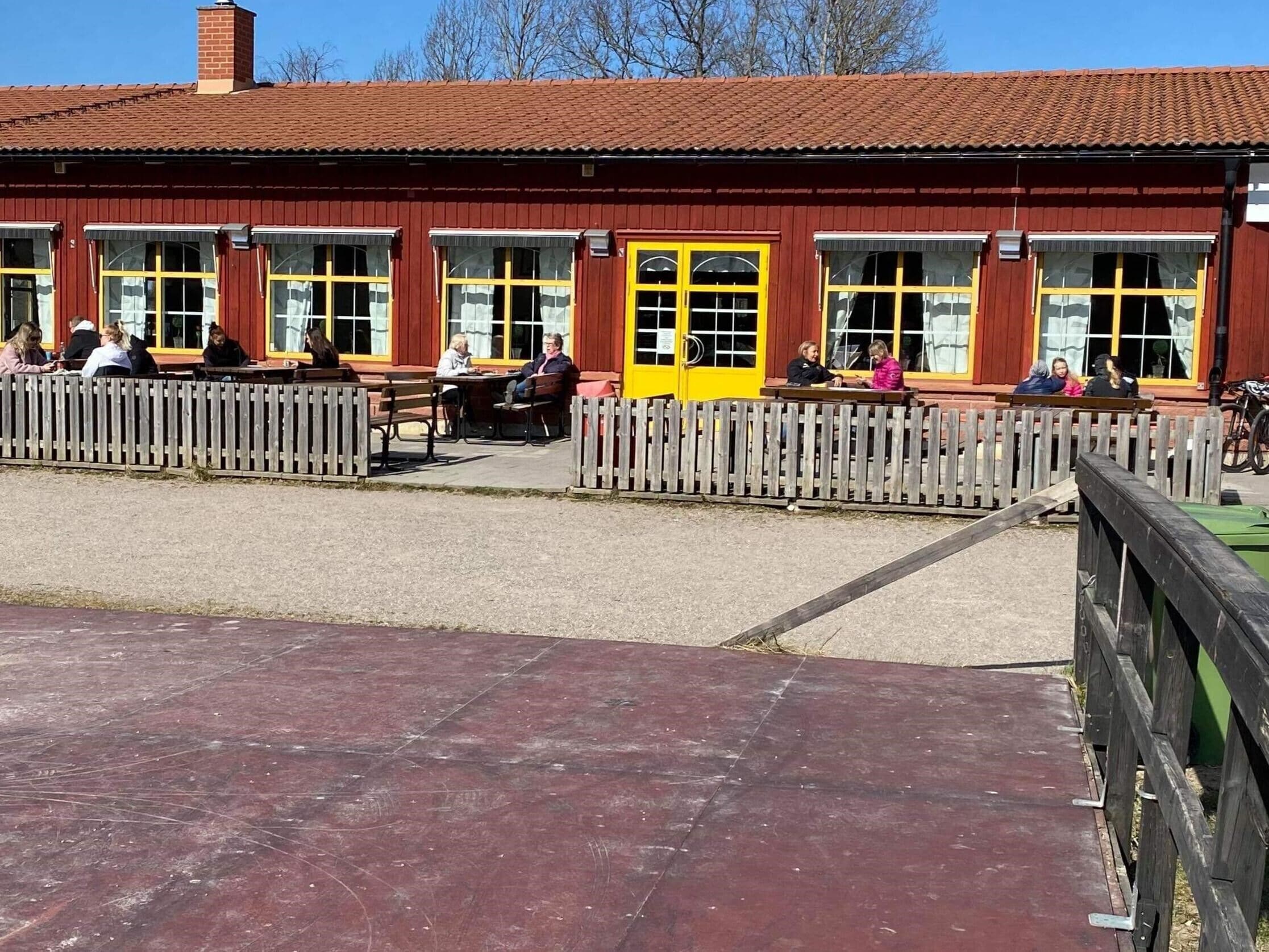 A red building with yellow-framed windows and a yellow door, featuring outdoor seating and a wooden fence.
