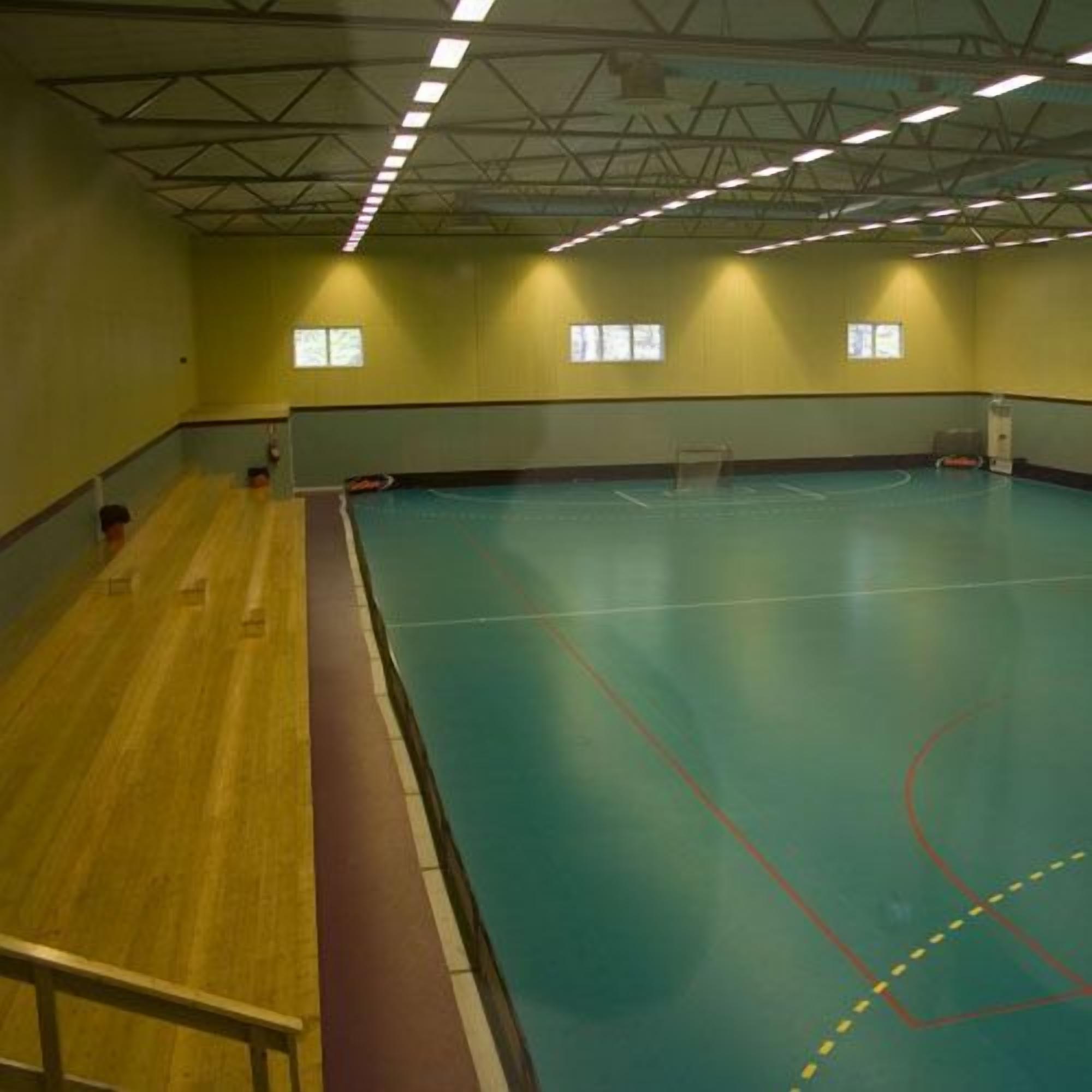 An indoor sports hall with a basketball court, seating area, and windows.