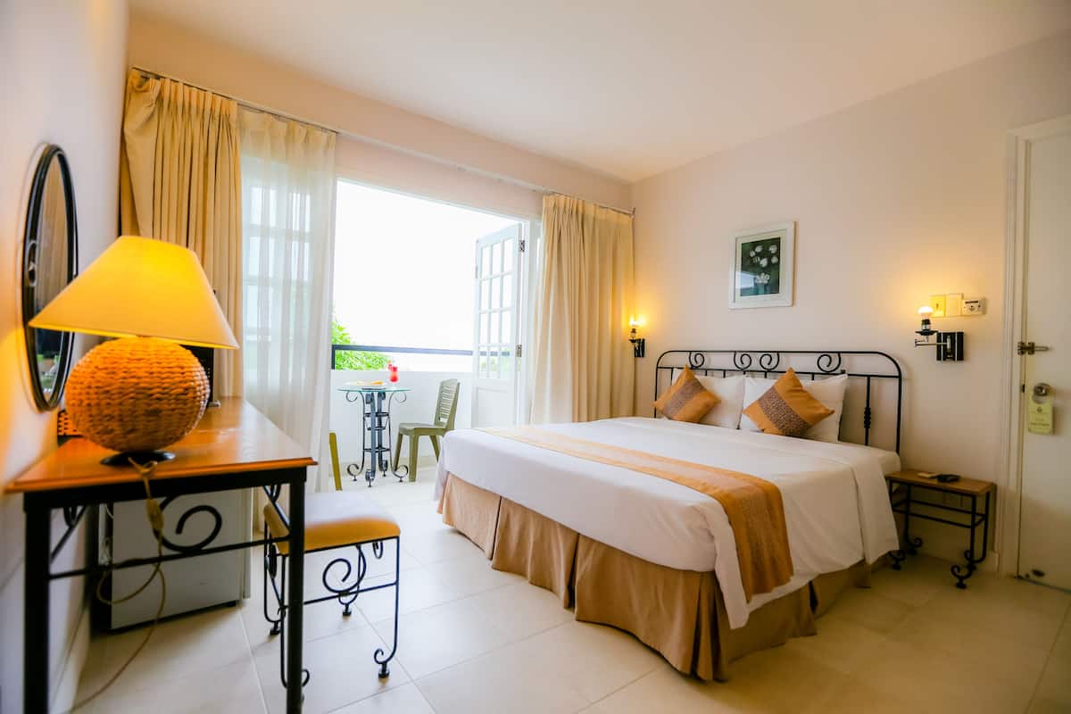 A hotel room with a bed, a desk with a lamp, a chair, a small table, and a view of the outdoors.