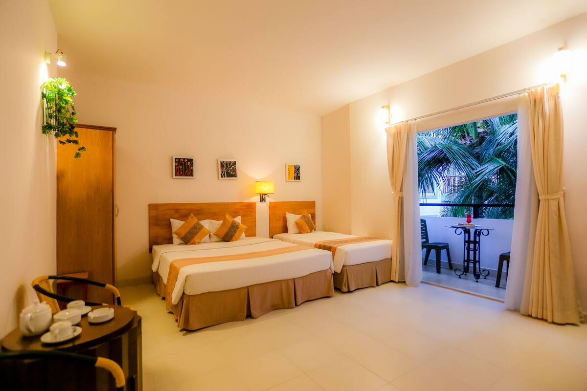 A hotel room with two beds, a small table with tea set, a balcony with a table and chairs, and a view of palm trees.
