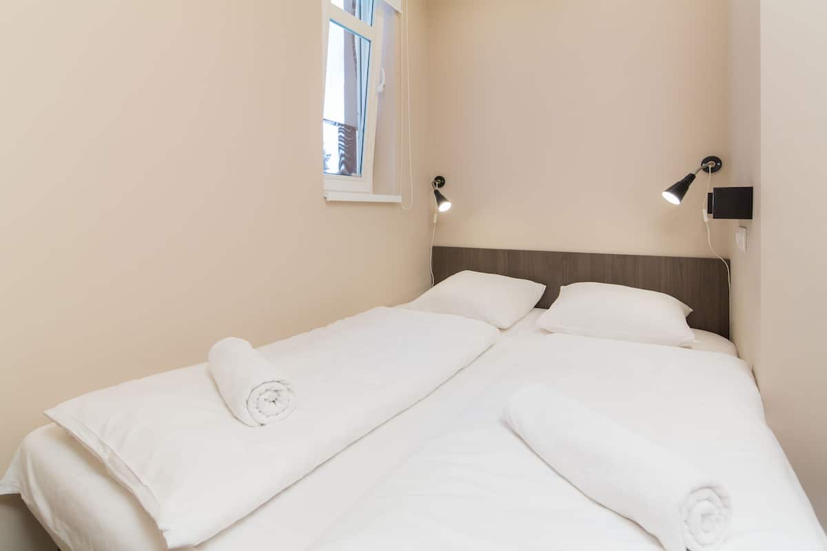 A neatly made twin bed with white linens and rolled towels, a bedside table with a lamp, and a window with curtains.