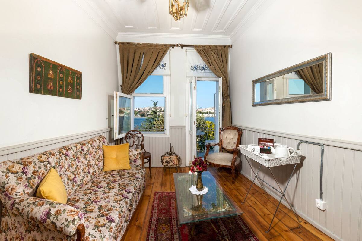 A cozy living room with a floral sofa, a coffee table, a chair, a small table with a basket, and a view of the sea through open curtains.