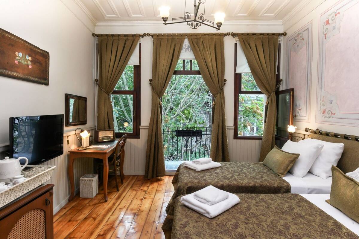 A hotel room with two beds, a desk with a TV, a small table with a lamp, and a view of trees through large windows.