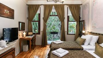 A hotel room with two beds, a desk with a TV, a small table with a lamp, and a view of trees through large windows.
