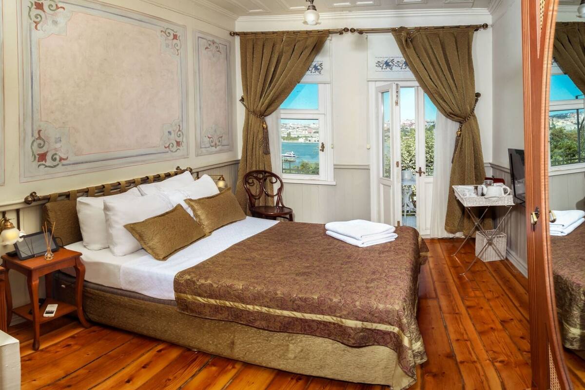 A bedroom with a large bed, a nightstand, a table with a kettle, and a view of the sea through the window.