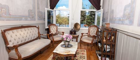 A room with a wooden floor, a sofa, two chairs, a table with a vase of flowers, a gramophone, and a bookshelf.