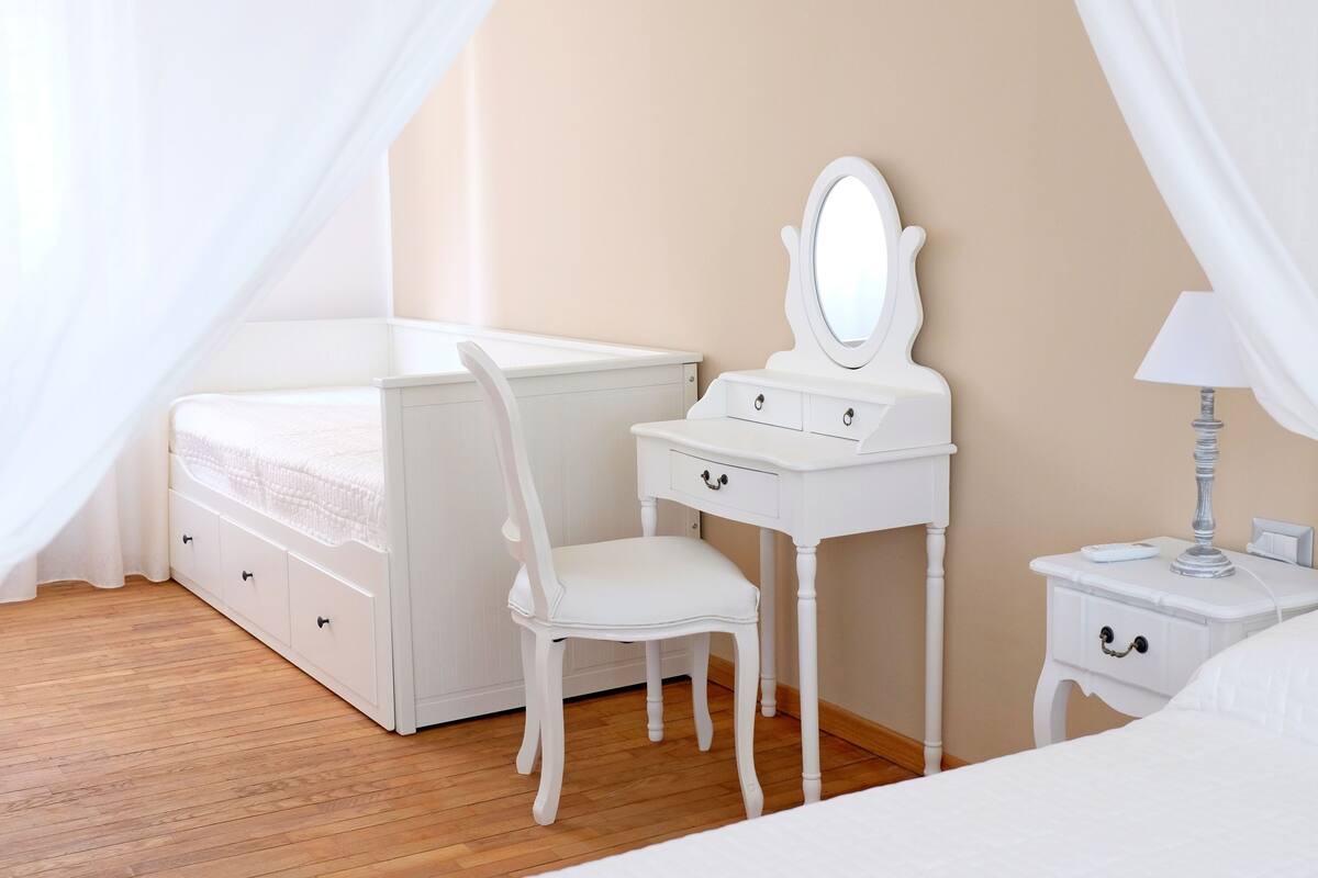 A bedroom with a bed, a vanity with a mirror, a chair, and a nightstand with a lamp.