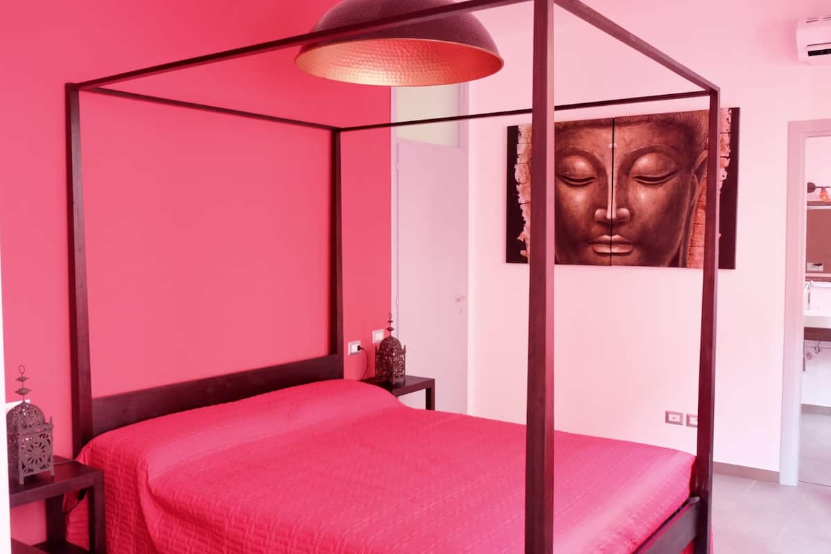 A room with a four-poster bed, a large Buddha painting, a modern air conditioning unit, and a bathroom visible through an open door.