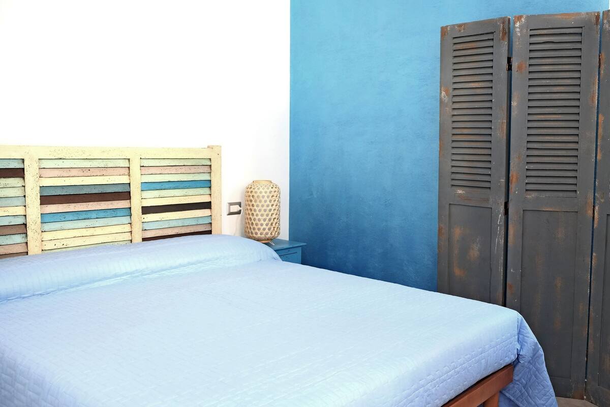 A room with a bed, a wardrobe, a bedside table, and a wall with a circular mirror.