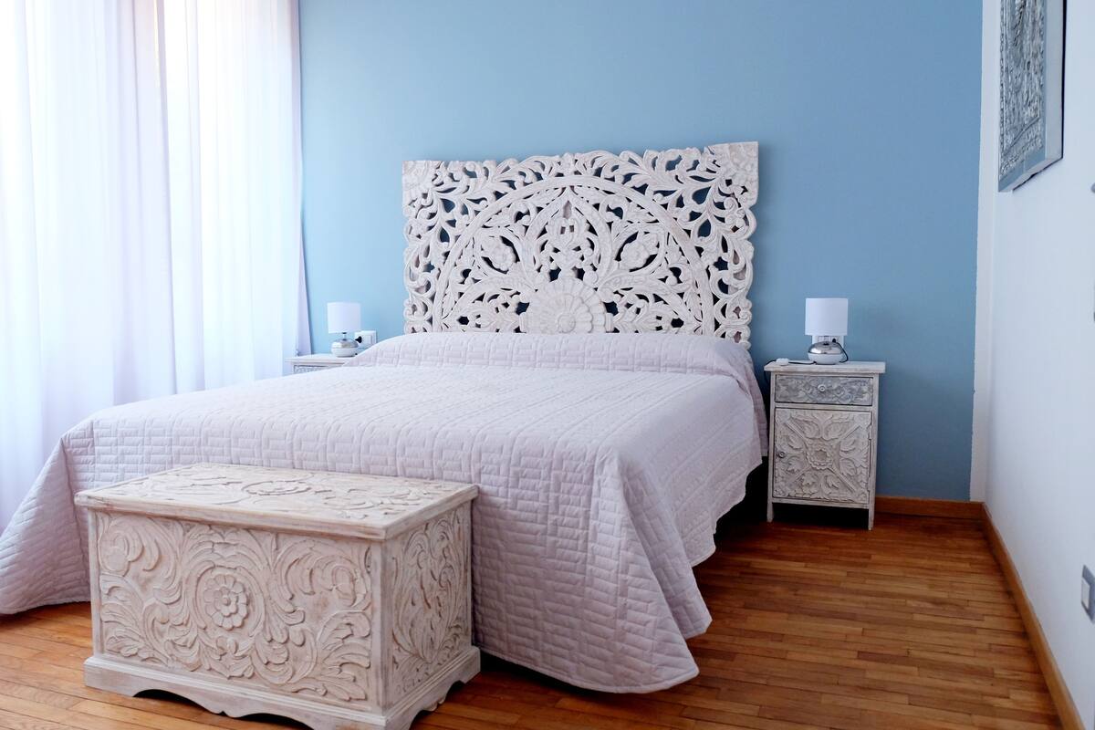 A bedroom with a bed, nightstands, a bedside table, and a decorative ottoman.