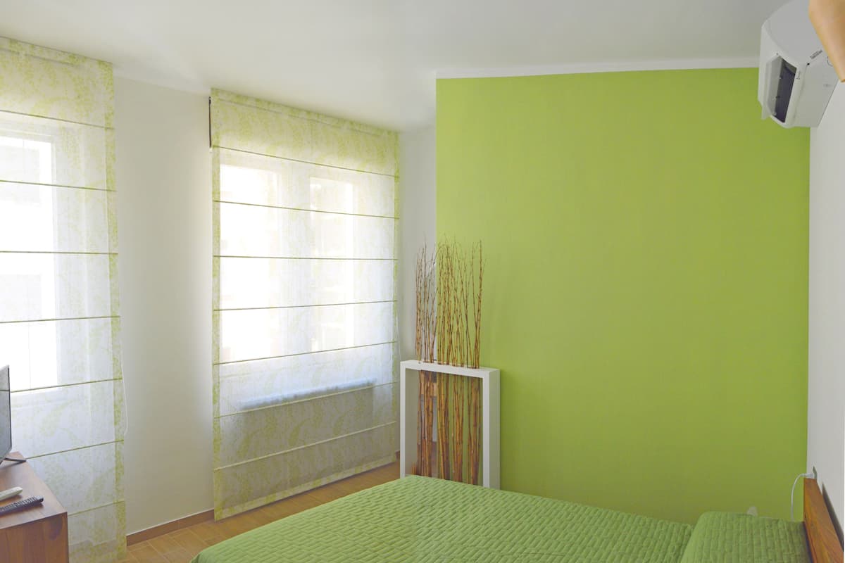 A bedroom with a green bedspread, a bedside table with a remote control, a window with a patterned curtain, and a wall-mounted air conditioning unit.
