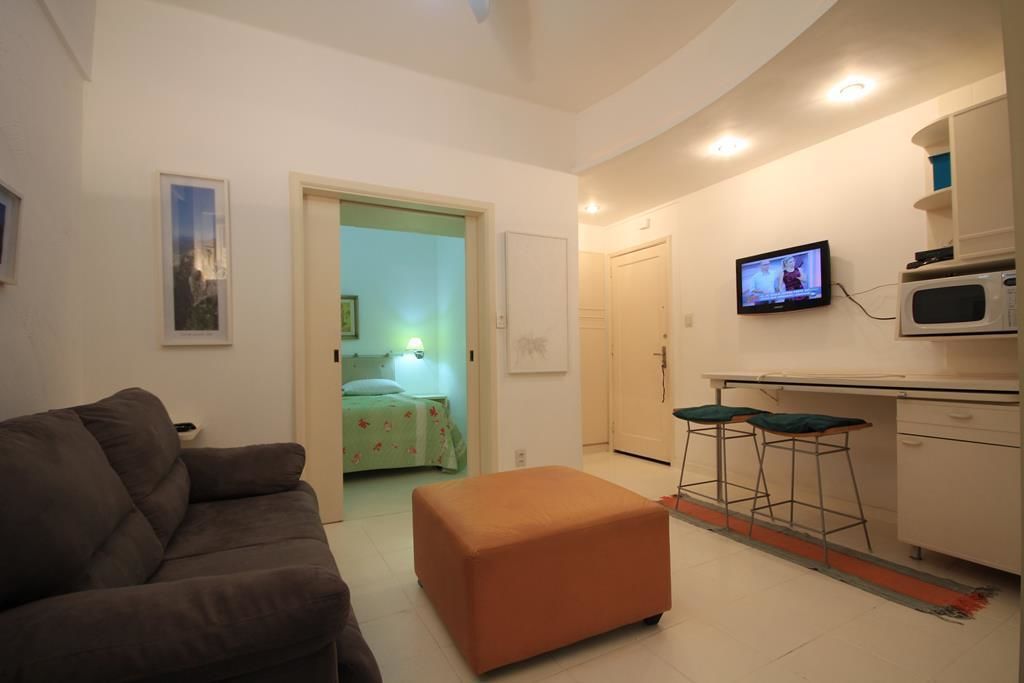 A compact living space with a sofa, an ottoman, a kitchenette with a microwave, a TV, and a bed in the adjacent room.