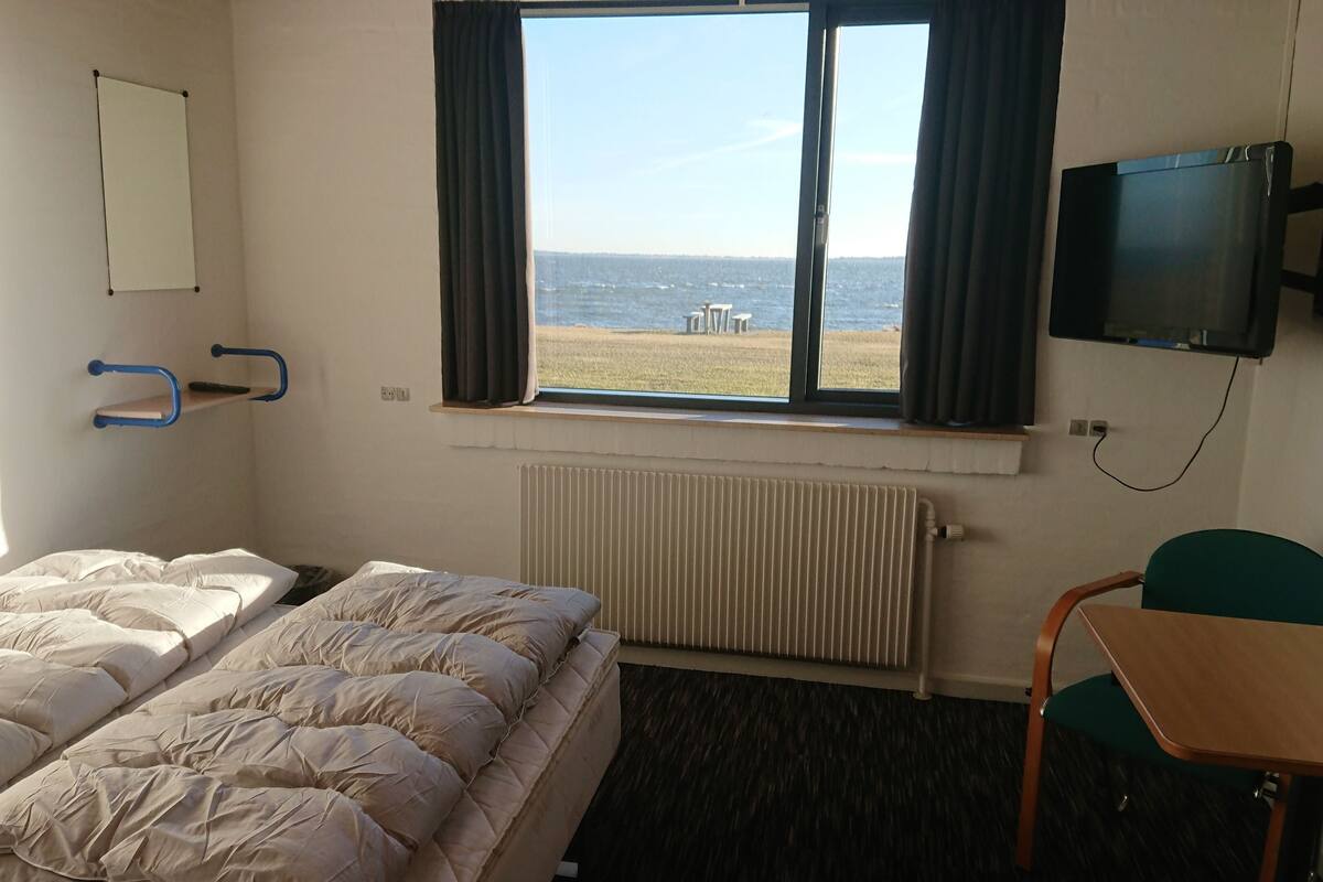 Een hotelkamer met een bed, bureau, stoel, televisie, raam met uitzicht op zee en een radiator.