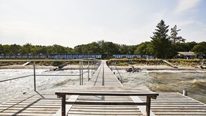 Un muelle de madera que lleva a un edificio con una hilera de ventanas, rodeado de árboles y una playa de arena.