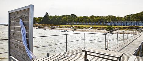 A wooden deck with benches overlooking a beach, a wooden board with towels, and a row of beach huts in the background.