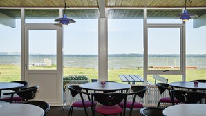Un área de estar interior con mesas y sillas, ventanas amplias con vista al mar y un techo de madera.