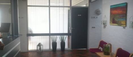 Una habitación con suelo de ladrillo, puerta corredera de vidrio, escritorio con computadora, dos plantas en macetas, una silla roja y un cuadro en la pared.