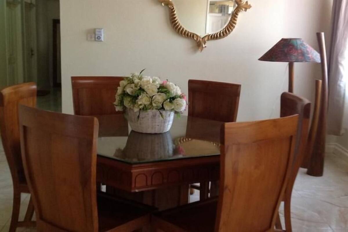 Un comedor con una mesa redonda, seis sillas de madera y un florero con flores blancas.