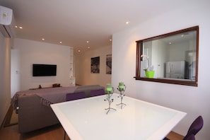 A modern, well-lit room with a bed, a white table with two green candle holders, a flat-screen TV mounted on the wall, and a window with a view of the kitchen. - Senna 1804 (Rio de Janeiro)