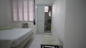 A small, clean room with a bed, a small table, a window with blinds, a toilet, and a sink. - Ferreira 201 (Rio de Janeiro)