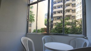 Ein Zimmer mit einem kleinen weißen Tisch und vier weißen Plastikstühlen. Ein großes Fenster bietet Blick auf ein mehrgeschossiges Gebäude und Grünflächen.